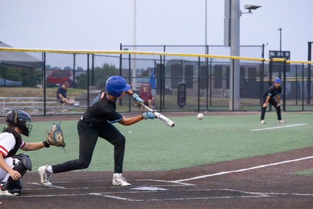 A young baseball player in a blue helmet swings at a pitch on a baseball field, while a catcher in a black helmet prepares to catch the ball.