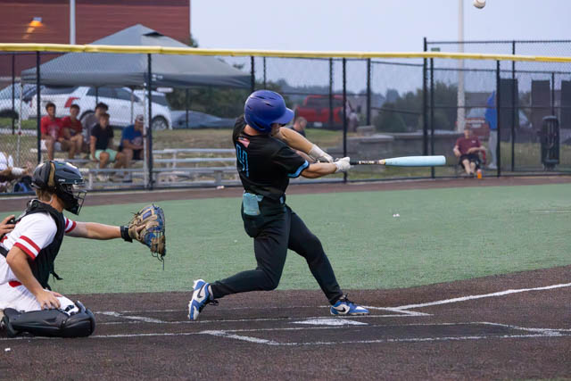 A baseball player in a blue helmet swings a bat to hit a ball, while a catcher in black gear crouches behind home plate during an evening game.