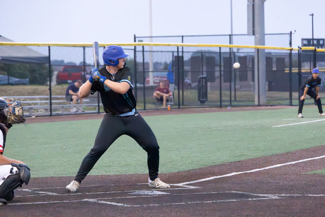 A young baseball player in a blue helmet and black uniform is about to hit a baseball with a bat while a catcher and another player are positioned nearby.