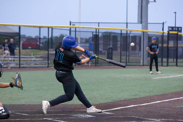 A young baseball player in a black uniform with the number 11 swings a bat at a pitched ball during a game on a field. Another player crouches behind home plate, while teammates are positioned in the background.