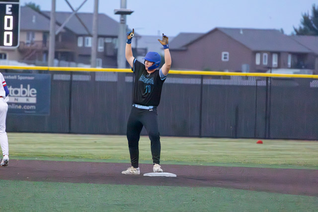 A baseball player in a black uniform with blue accents celebrating on second base with hands raised in the air.