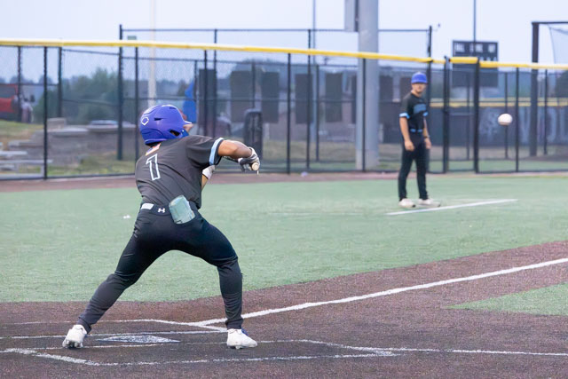 A young baseball player in a blue helmet and grey uniform prepares to hit a pitch during a game, while another player stands ready to pitch in the background.