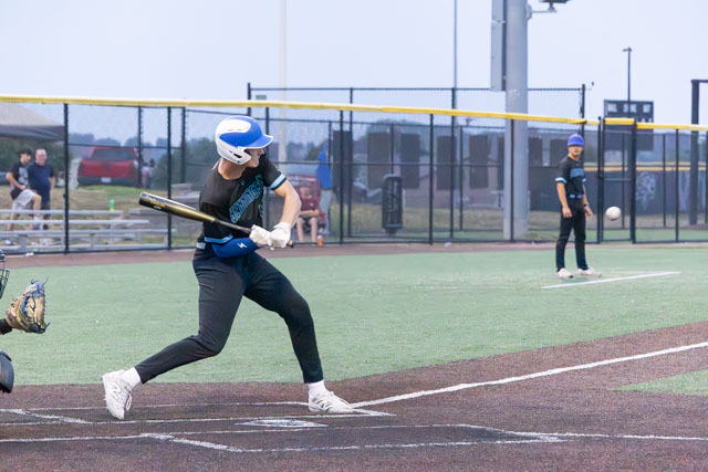 A young baseball player swinging a bat at home plate during a game, with a catcher in the foreground and a pitcher in the background.