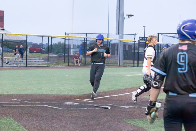 A baseball player in a dark uniform with a blue helmet runs towards home plate while another player in a white uniform plays catcher. A bat lies on the ground near home plate.