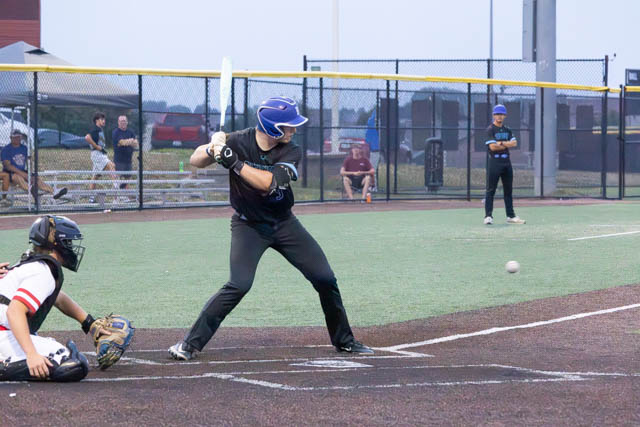 A baseball player in a blue helmet and black jersey swings at a ball while a catcher in a white jersey crouches behind home plate. In the background, spectators are watching the game.