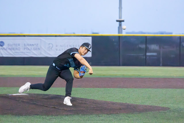 A young baseball pitcher in a black uniform throws a pitch from the mound during a game.