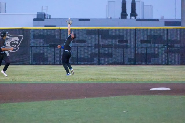 A baseball player leaping to catch a ball in the outfield during a game, with a teammate running in the background.