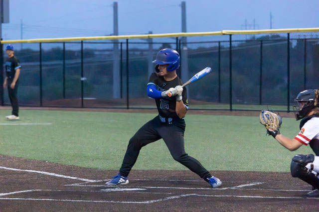 A young baseball player in a black uniform with blue accents prepares to bat, with a catcher in a white and black uniform ready behind home plate.