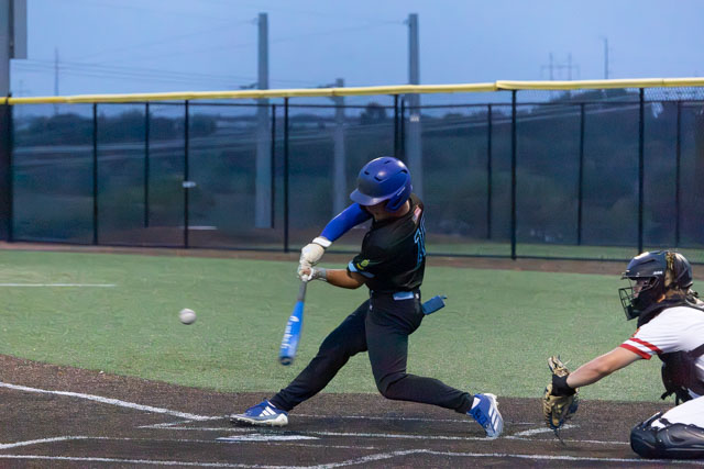 A baseball player in a blue helmet and jersey swings a bat at an approaching ball while a catcher in a black helmet and shirt crouches behind home plate, set against a twilight sky.