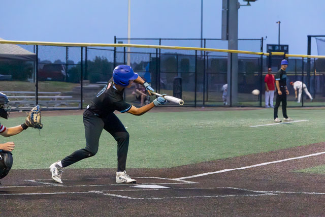 A baseball player in a blue helmet is swinging a bat, attempting to hit a pitched ball on a well-maintained field during a game. Other players are visible in the background.