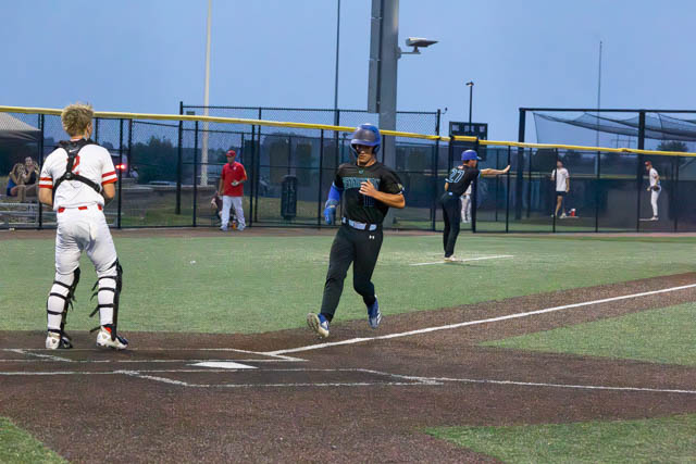 A player in a black uniform runs towards home plate while a catcher in a white uniform prepares to receive the ball. Other players are visible in the background on a baseball field during dusk.