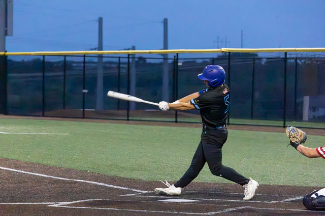 A baseball player swings a bat while standing at home plate during a game at dusk.