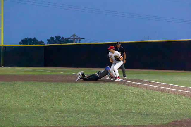 A baseball player in a red cap and white uniform reaches for the ball at first base while a player in a black uniform slides toward the base. An umpire watches the play.