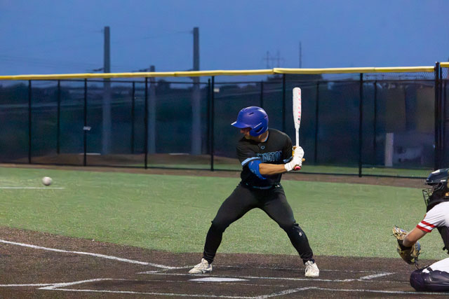 A baseball player in a blue helmet and black jersey prepares to bat as a ball approaches during a night game.