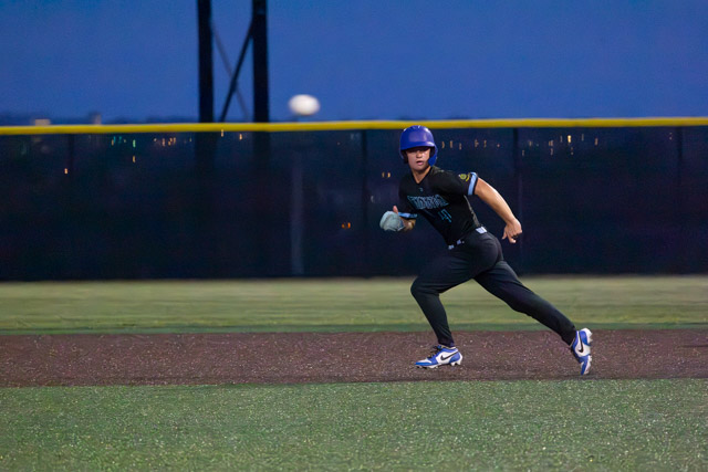 A young baseball player in a blue helmet and black uniform running on a baseball field under the evening sky.