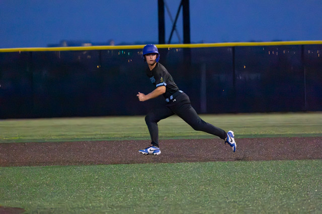 A player wearing a blue helmet and a black uniform running on a baseball field at dusk.