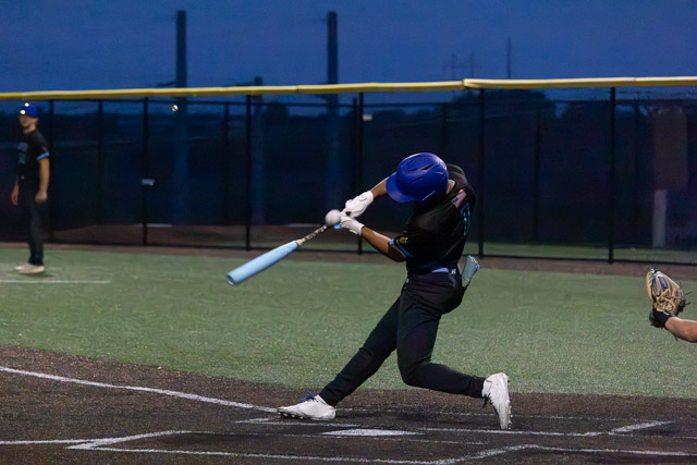 A baseball player in a blue helmet swings a bat at home plate during a night game, with the field illuminated by stadium lights.