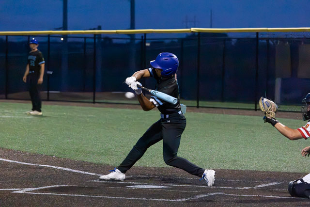 A baseball player swings a bat at a pitch during a night game, with a catcher positioned behind home plate.