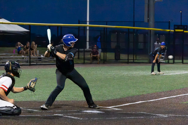 A baseball player in a blue helmet and uniform prepares to hit the ball at home plate, while the catcher squats behind him and the pitcher is in the background ready to throw.