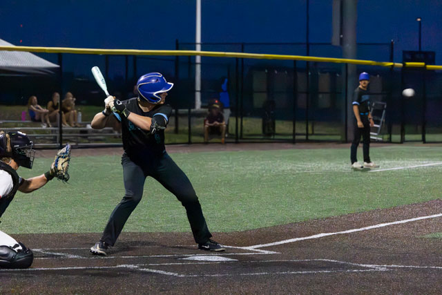 A baseball player is in the batting stance, poised to hit a ball coming toward him. The player is wearing a blue helmet and black uniform, standing in a baseball field at dusk with spectators in the background.