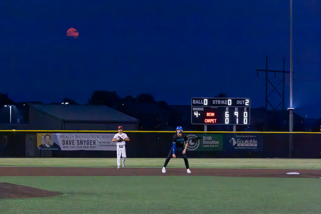 A baseball field at night with a scoreboard displaying the inning and score, two players in position, and a red moon visible in the background.