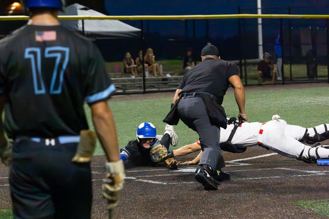A baseball player in a blue helmet dives towards home plate while a catcher in white gear prepares to tag him out, with an umpire in the foreground overseeing the play.