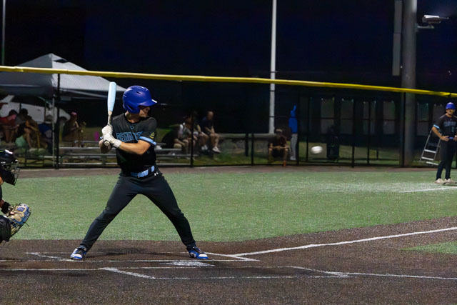 A baseball player stands at home plate, poised to bat, with a catcher in position. The game is being played at night on a well-lit field.