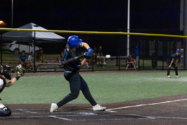 A baseball player in a blue helmet swings a bat at home plate during a night game, with a catcher in gear and spectators in the background.