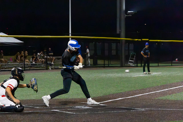 A baseball player in a blue helmet and black uniform swings at a pitch during a night game, while a catcher in a black and red uniform crouches behind home plate.