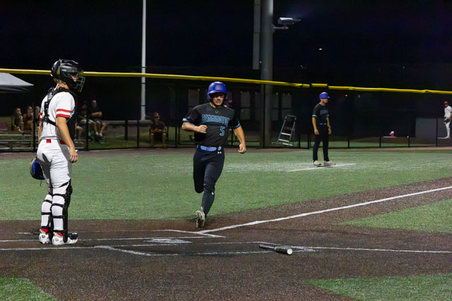 A baseball player in a dark uniform and helmet joyfully runs towards home plate, while a catcher in a black and white uniform watches. The scene is set on a well-lit field during a night game.