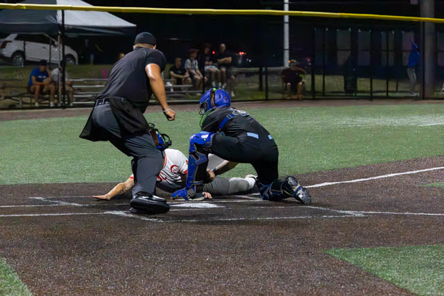A baseball game scene at night, featuring a catcher in blue gear attempting to tag a sliding player at home plate, with an umpire in black nearby and spectators in the background.