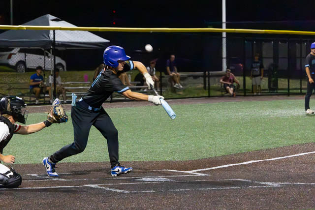 A baseball player in a blue helmet swings at a pitch during a night game, with the catcher ready to receive the ball.