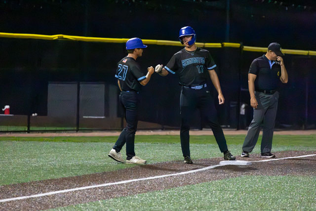 Two baseball players celebrating at home plate during a game, with one player wearing a blue helmet and jersey number 17, and the other wearing jersey number 27. A coach or umpire is visible in the background.