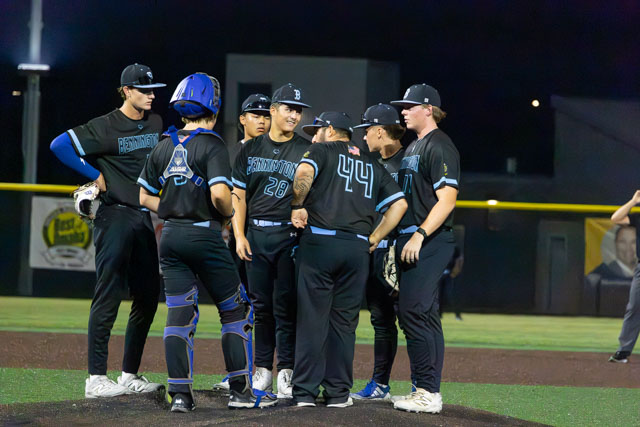 A group of baseball players in black uniforms huddle together on the field at night, discussing strategies.