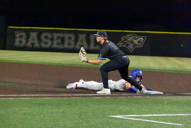 A baseball player crouches at first base while a runner slides towards the base.