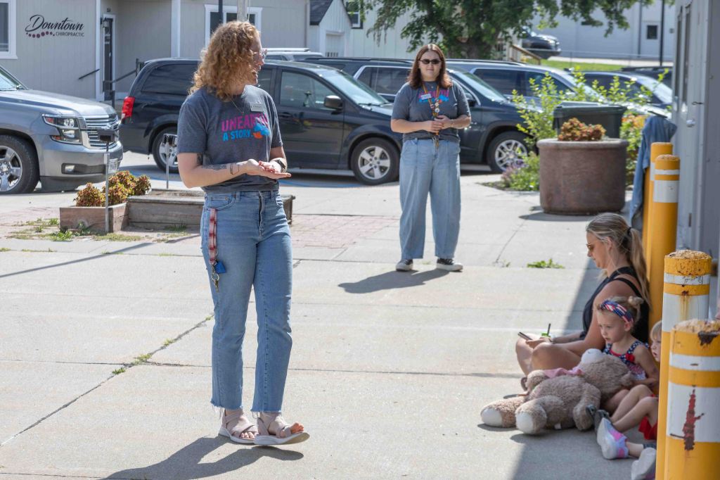 A group of people outside, with one woman standing and holding a phone, while another is seated with children and stuffed animals, in a sunny urban setting.