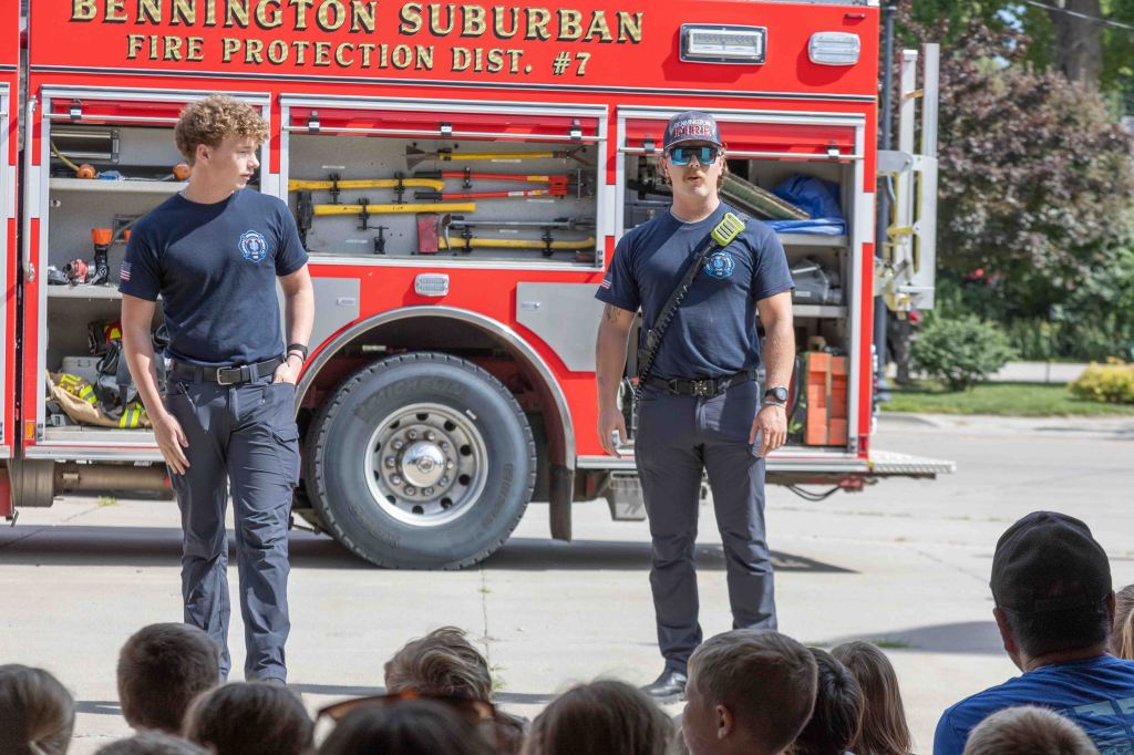 Two firefighters standing in front of a fire truck, addressing a group of children seated outside.
