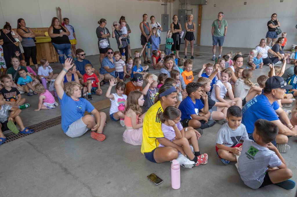 A large group of children and adults gathered in a communal space, with many children raising their hands to participate. The environment appears lively and engaging, with adults observing and facilitating.