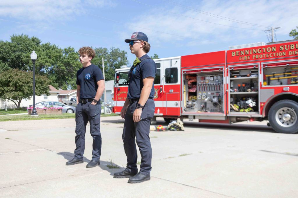 Two firefighters standing in front of a red fire truck with equipment visible, on a clear day.