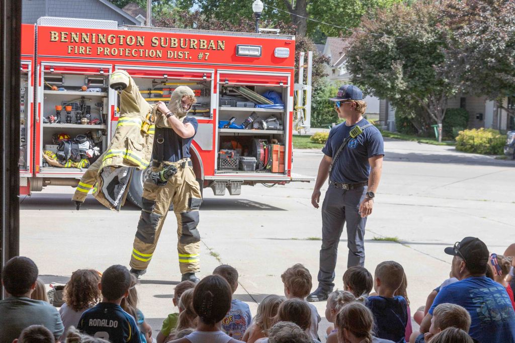 A firefighter demonstrating how to put on firefighting gear in front of a group of children, with a fire truck in the background.