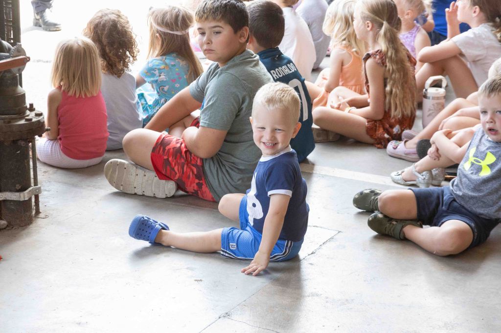 A group of children sitting on the floor, some looking towards the front, with a smiling young boy in a blue outfit sitting in the foreground.