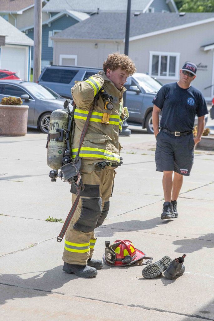 A young firefighter in a full protective uniform stands on a concrete surface, adjusting his equipment, while another firefighter in casual attire observes nearby. A helmet and boots are placed on the ground in front of them.