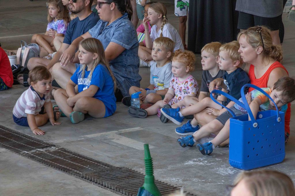 A group of children and adults sitting together on the floor, engaged in an activity, with some children watching intently and others playing.