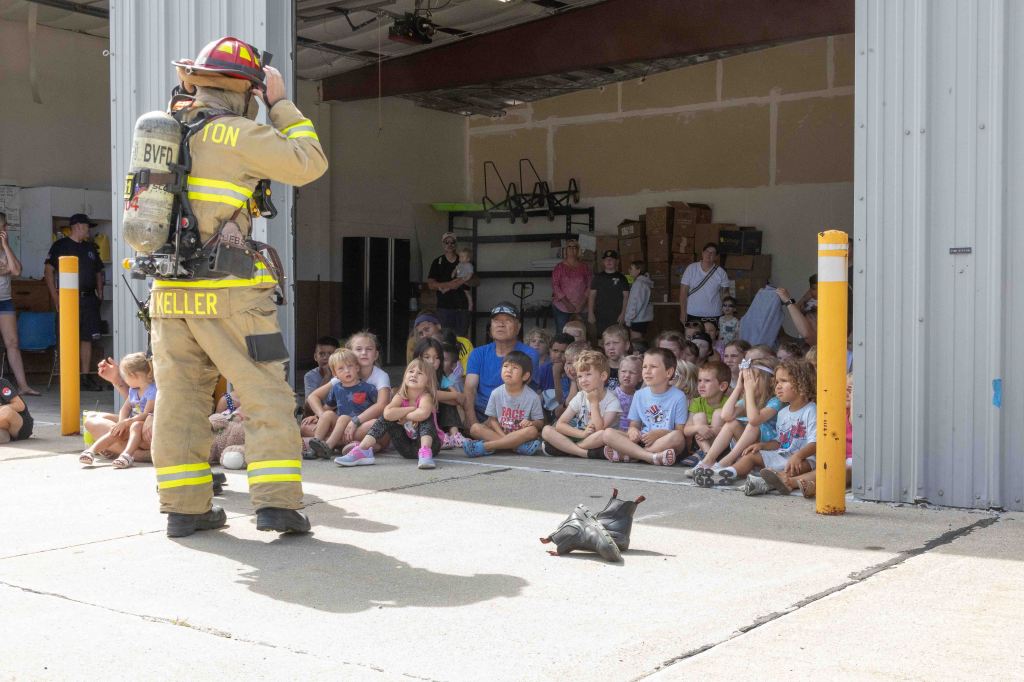 A firefighter saluting while addressing a crowd of children sitting on the ground, with adults observing in the background. The setting appears to be inside a garage or fire station.