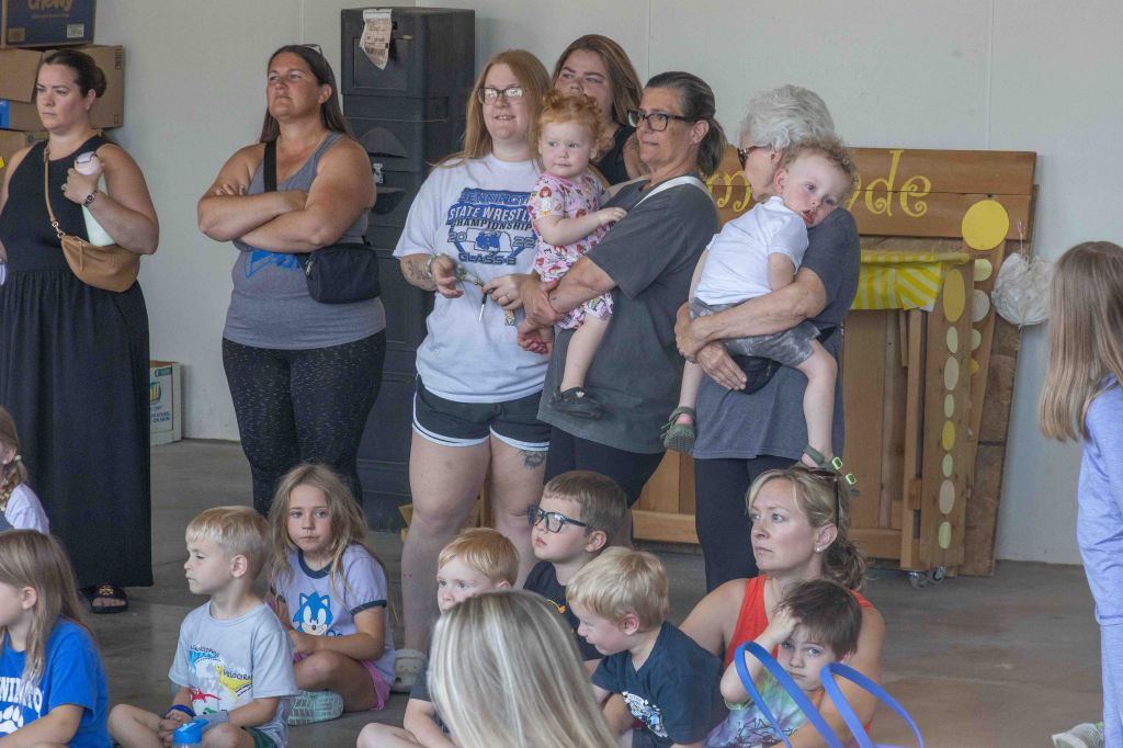 A group of adults and children gathered in a large indoor space, with some adults standing and watching while children sit on the floor. Several individuals hold young children, and there is a wooden sign in the background with yellow decorations.