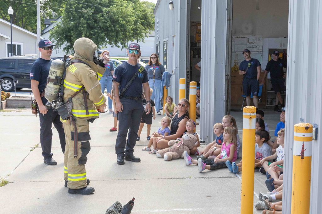 A firefighter in full gear demonstrates safety measures to a group of children sitting on the ground, accompanied by adults, outside a fire station.