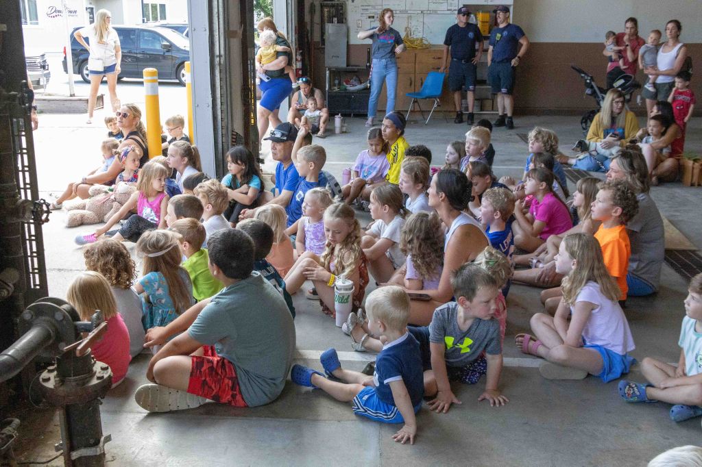 A large group of children sitting on the floor, attentively watching a presentation in a fire station, with adults in the background.