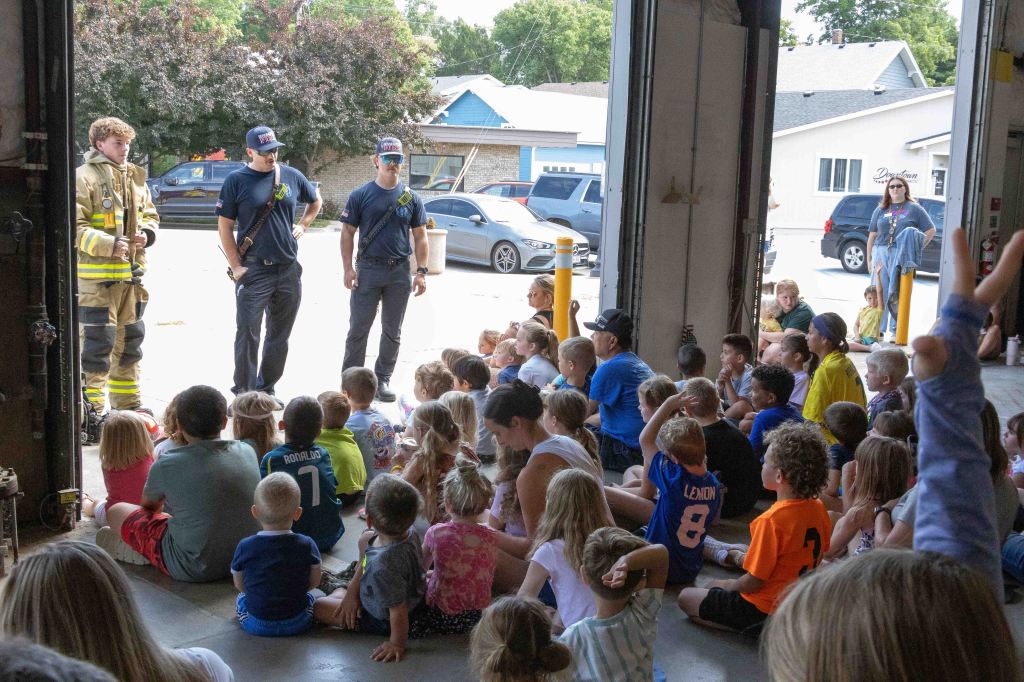A group of children sitting on the floor of a fire station, listening attentively to firefighters and staff. Two firefighters, one in full gear, stand alongside another firefighter in casual attire, addressing the kids with a vehicle visible in the background.