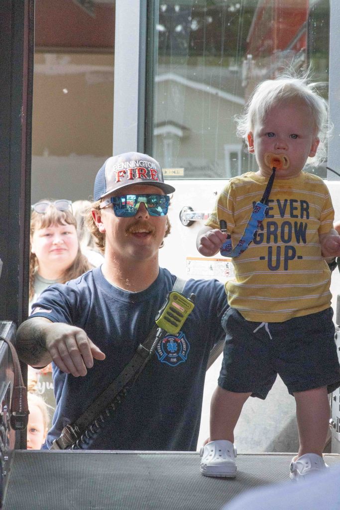 A firefighter in a blue shirt and cap interacts with a smiling toddler wearing a yellow shirt with the words 'NEVER GROW UP' and holding a pacifier, as they stand at the entrance of a fire truck.