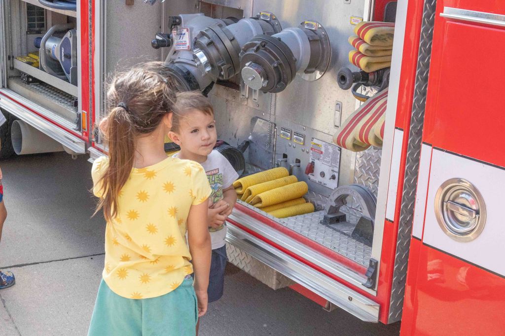 Two young children stand in front of a fire truck, interacting near the fire hose compartment.
